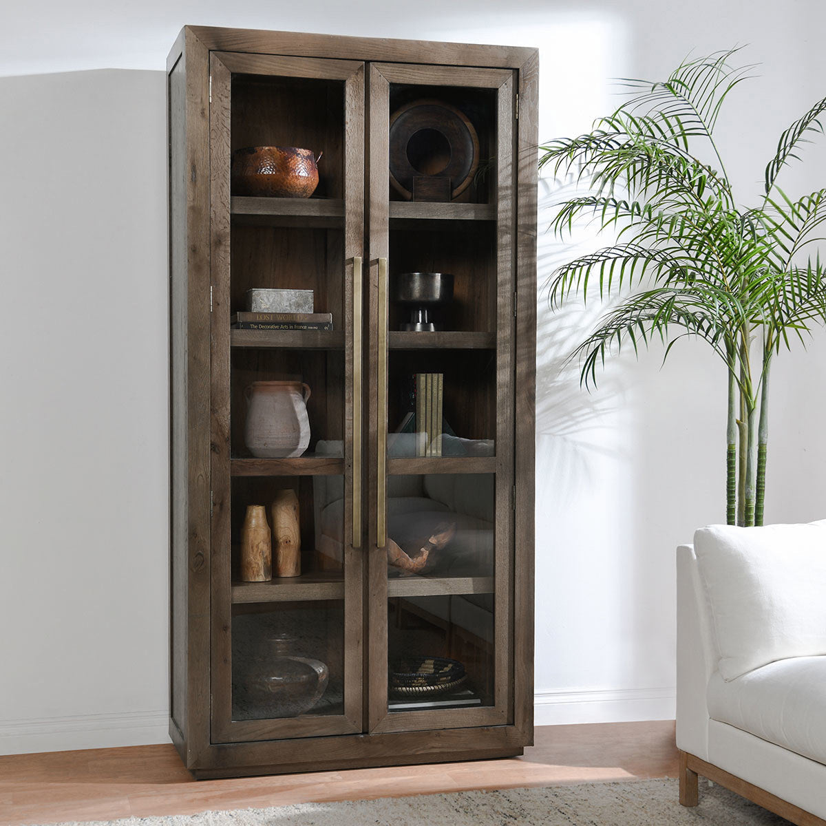 Tall glass door curio cabinet styled with books, ceramics, greenery, and living room seating