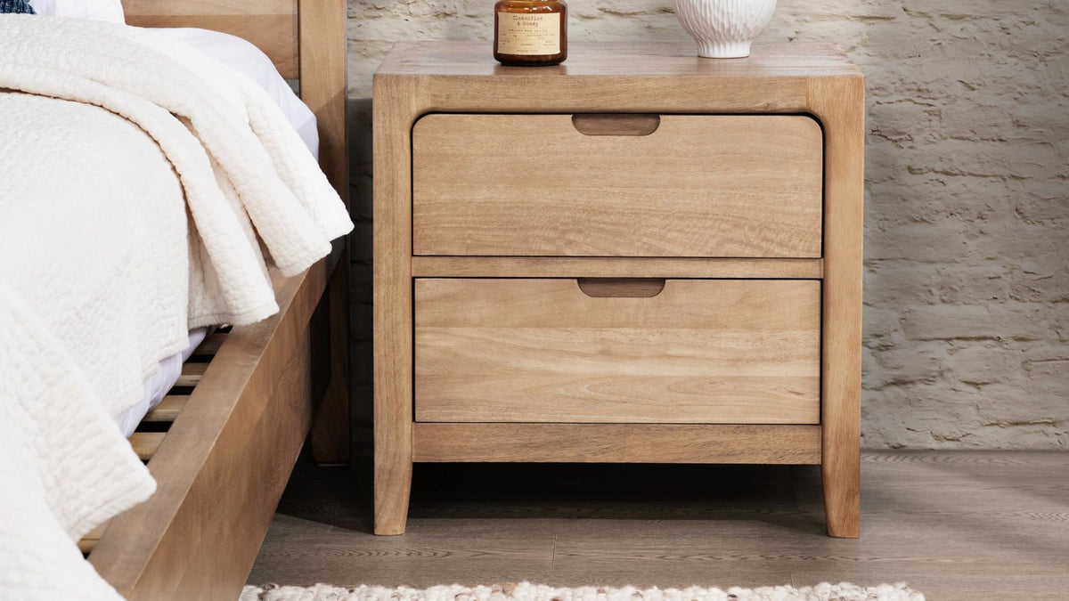 Natural wood two-drawer nightstand beside a coordinating bed in a softly styled bedroom