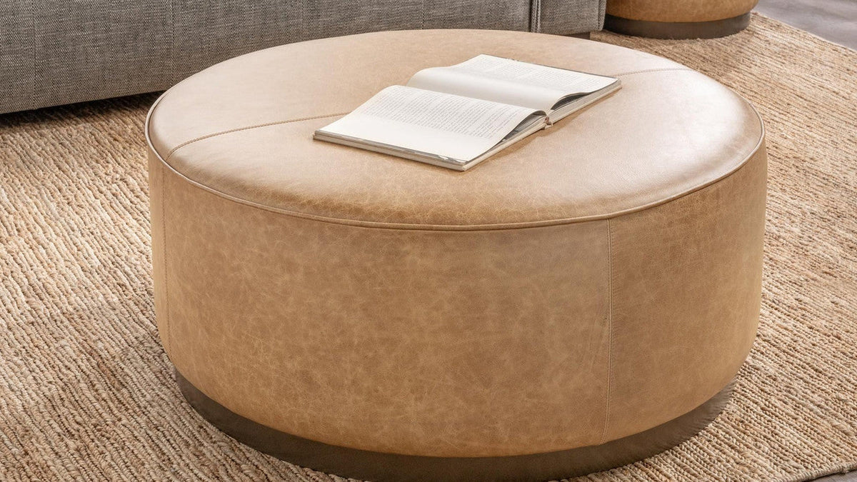 Round chestnut leather ottoman styled as a coffee table with a book beside a neutral sofa